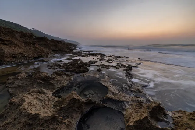 Shallow water lying in the sculpted rock pools of the shore platform at dawn, with the sea beyond and a forested headland to the left.