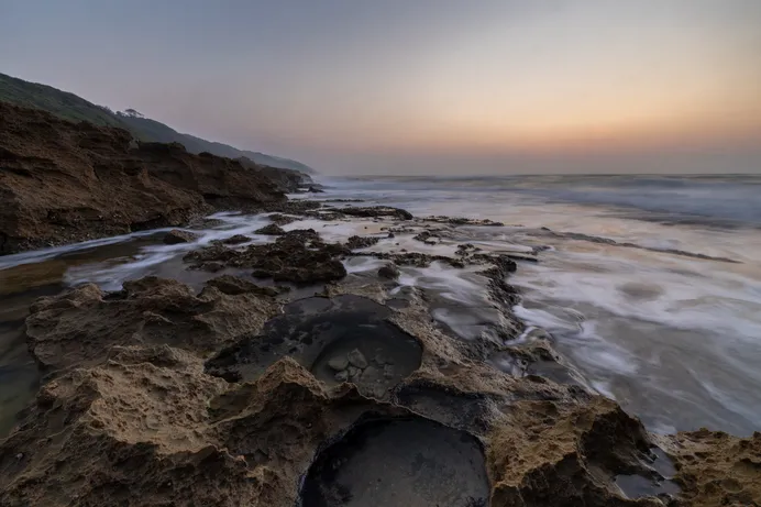 Sculpted rock pools in the sandstone shore platform at low tide, with the incoming surf blurred by a long exposure and a forested headland behind.