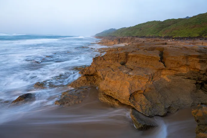 A weathered sandstone point on the rocky shore with turquoise surf washing around it and a green forested headland in the distance.