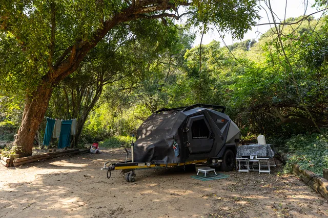 An off-road camping trailer set up on a sandy campsite under a large spreading tree, with towels hung on a line and a camp table and chairs beside it.